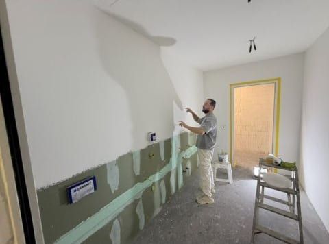 Ein Handwerker an einer oben weiss gestrichenen Wand, unten gespachtelte Plattenfugen