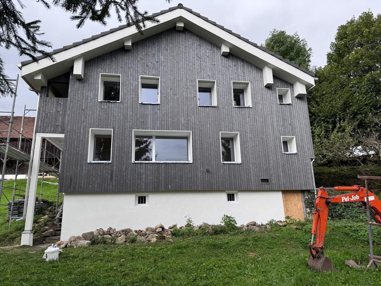 Haus mit dunkler Holzschalung, weissen Fensterumrandungen und weiss verputztem Sockel, daneben ein Gerüst