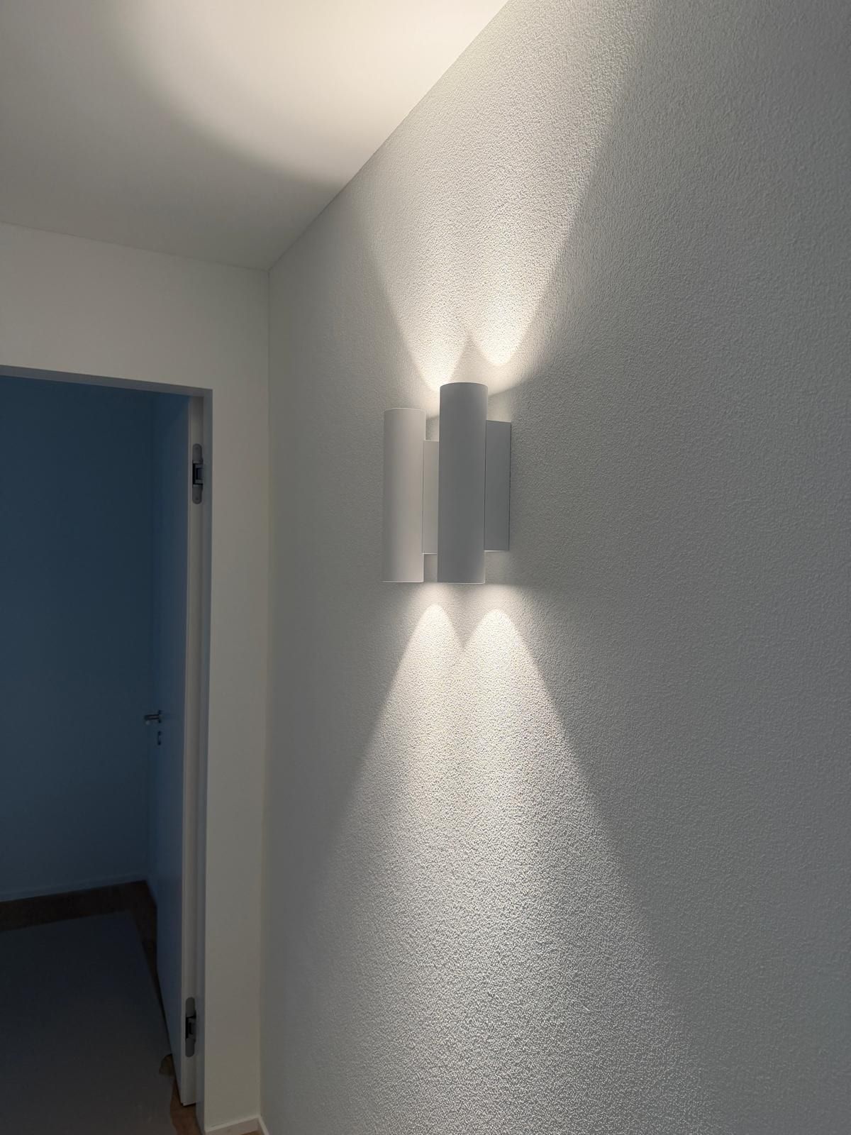 Wand mit Strukturputz, eine Wandleuchte wirft Lichtkegel nach oben und unten, daneben eine offene Tür