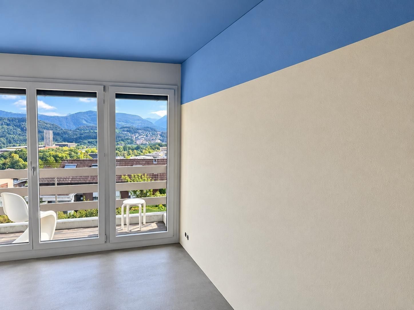 Raum mit blau gestrichener Decke und blauem Wandstreifen oben, darunter eine helle verputzte Wand, Fensterfront mit Blick auf Berge